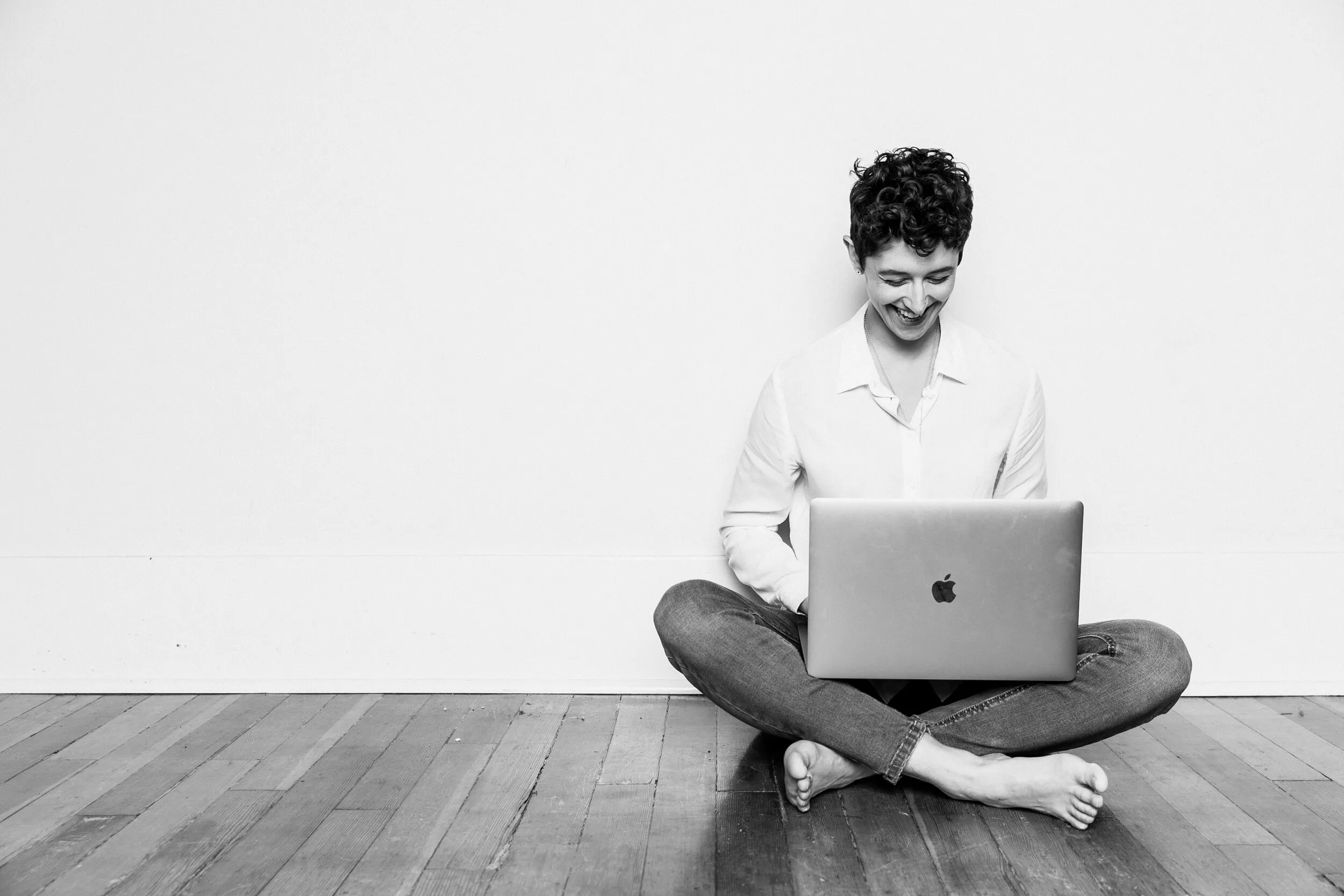 Gillian sitting cross-legged on a wooden floor, laughing, with a laptop on their lap