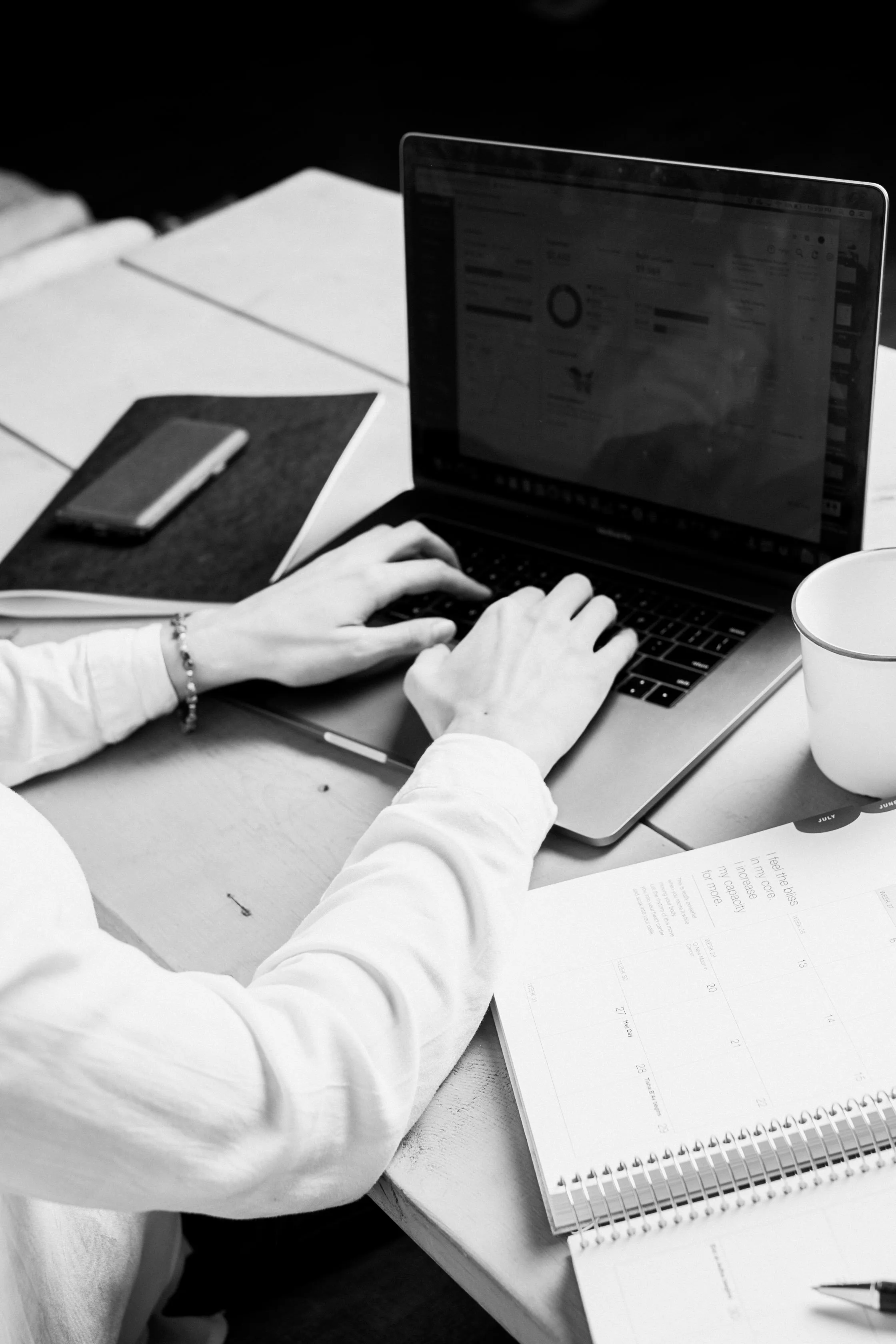 Hands typing on a laptop beside a notebook and a mug